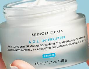 Frasco de crema antienvejecimiento A.G.E. Interrupter de SkinCeuticals, en una mano, sobre fondo azul claro.