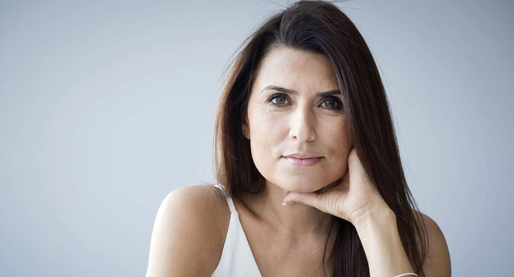 Mujer de cabello largo y oscuro, posando con una mano en la barbilla, mirada reflexiva y fondo claro.