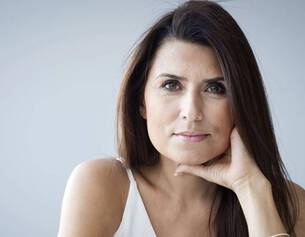 Mujer de cabello largo y oscuro, posando con una mano en la barbilla, mirada reflexiva y fondo claro.