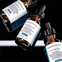 Tres frascos de serum con Vitamina C de SkinCeuticals, sobre un fondo oscuro y reflectante.