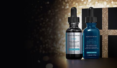 A close-up shot of two SkinCeuticals serum bottles, A.G.E Interrupter Ultra Serum and P-Tiox, standing in front of a dark gift box with a sparkling gold ribbon.