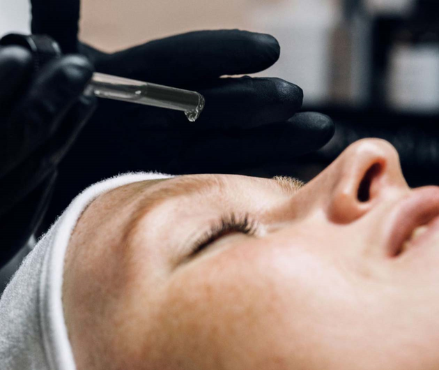 Una persona recibe un tratamiento facial, mientras un profesional aplica un serum con un gotero.