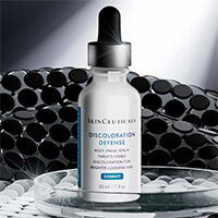 Un frasco de serum Discoloration Defense de SkinCeuticals sobre una superficie reflectante, contra un fondo oscuro y texturizado.​