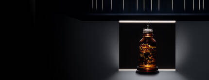 Botella de vidrio ambar iluminada desde abajo, situada en un entorno oscuro con detalles arquitectonicos en el fondo.