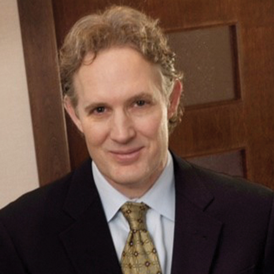 A professional man with curly hair and a suit, smiling in an office setting with wood paneling in the background.