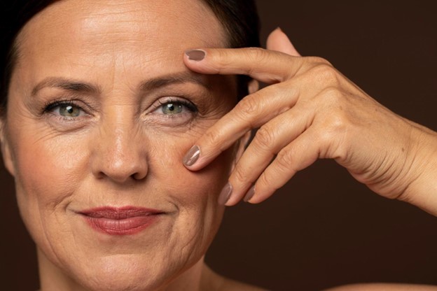 Mujer de mediana edad sonriente con piel radiante, tocándose suavemente la cara con una mano. Fondo marrón.