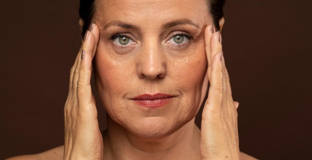 Mujer de mediana edad con expresión serena, sosteniendo su rostro con ambas manos sobre un fondo marrón.