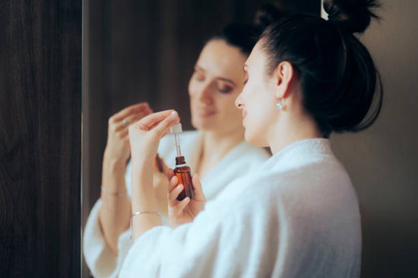 Mujer en bata de baño aplicando un suero facial frente al espejo, sonriendo con un cuidado personal.