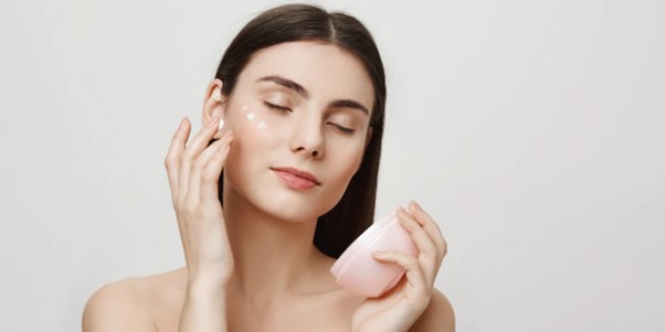 Mujer aplicando crema facial en su piel, sosteniendo un tarro de crema rosa, sonriendo suavemente.