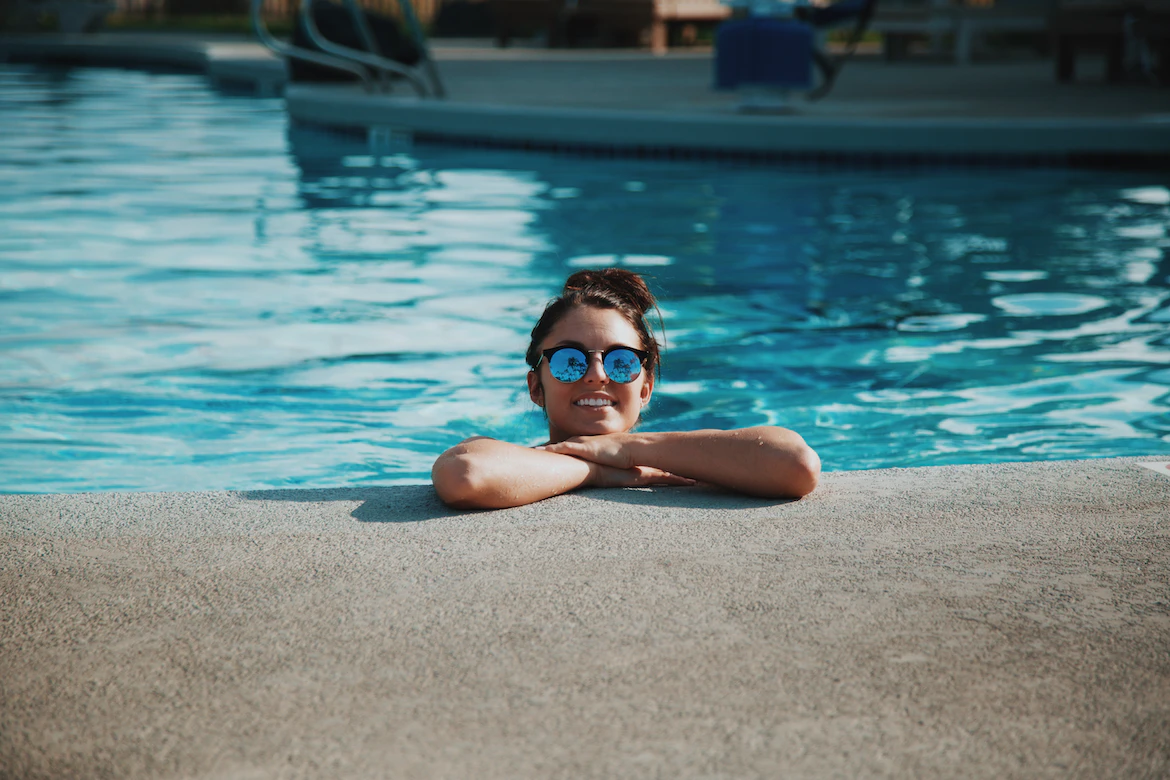 Una joven sonriente con gafas de sol se relaja en el borde de una piscina, con el agua cristalina reflejando el sol.