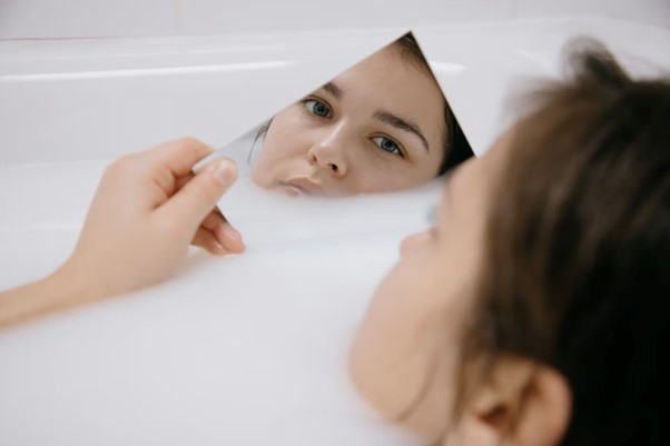 Una persona mira su reflejo en un espejo mientras está sumergida en una bañera llena de espuma.